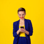 Happy young woman holding using new smartphone connected browsing internet worldwide isolated on yellow wall background. 4g data plan provider